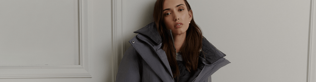 A woman wearing a dark gray oversized puffer jacket with a high collar, leaning against a white paneled wall.
