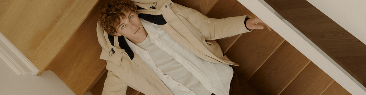 A man wearing a light beige trench coat over a white shirt, looking down the wooden stairs.