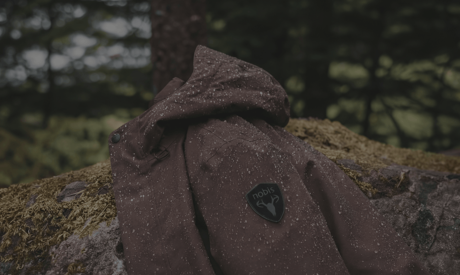 Nobis Upton Men's All-Weather Shell Jacket in Coffee Bean colour laying on a mossy rock in the forest.