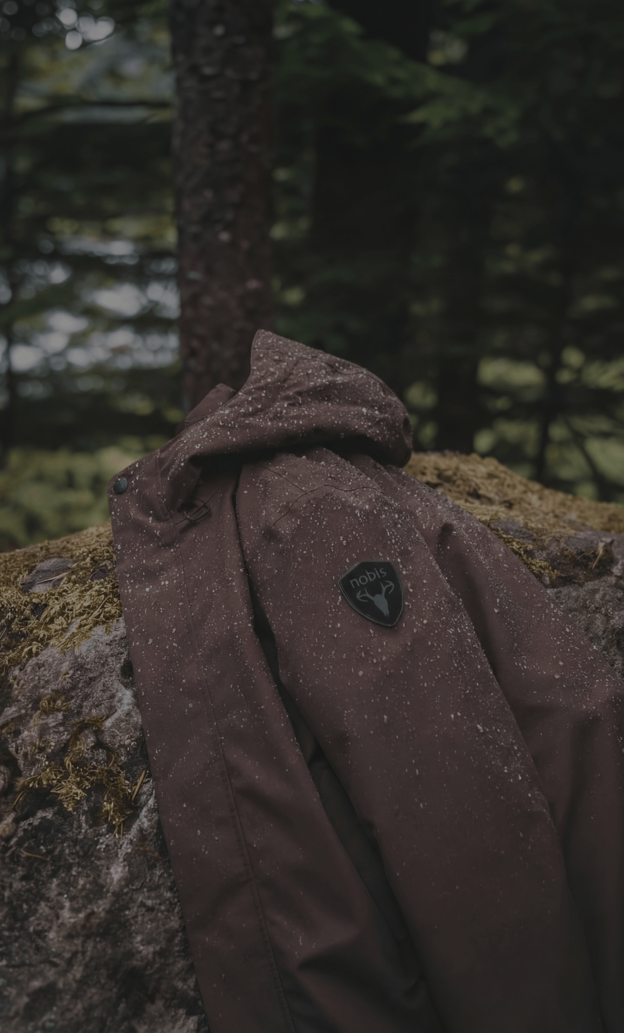 Nobis Upton Men's All-Weather Shell Jacket in Coffee Bean colour laying on a mossy rock in the forest.